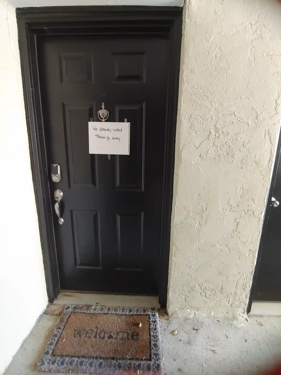 A black front door with a silver handle and digital lock has a handwritten note taped to it reading 'We already voted Please…