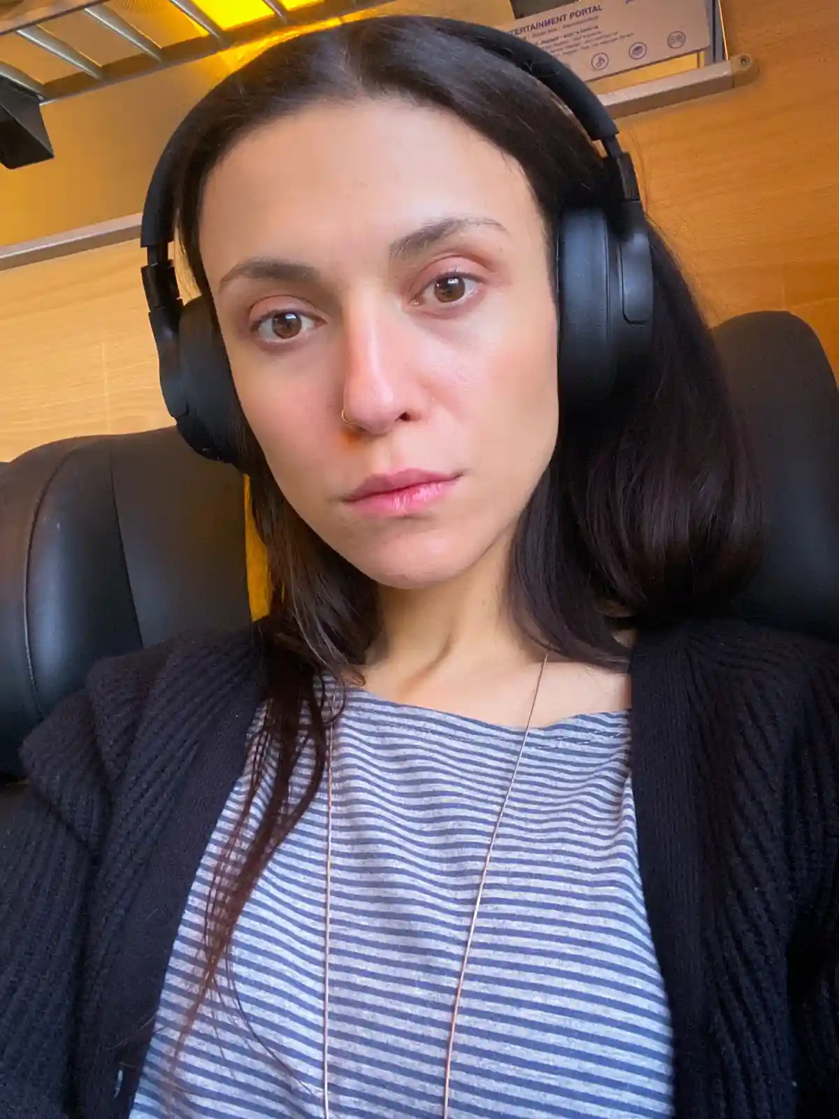 A woman with dark hair and a nose ring wears large black over-ear headphones while sitting in a seat with a wooden panel and…