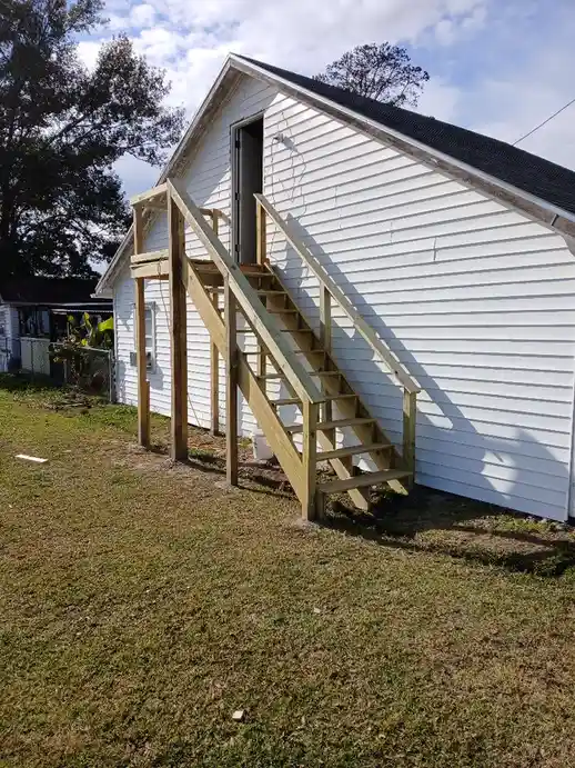 An exterior photo of a white house with a wooden staircase leading up to an upper-level door. The stairs are unfinished…