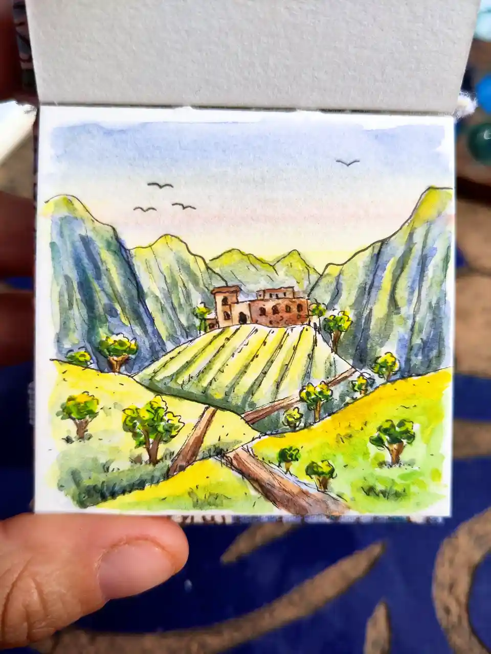 a hand holds a small watercolor painting depicting a scenic mountain landscape with a building on a hill, surrounded by…
