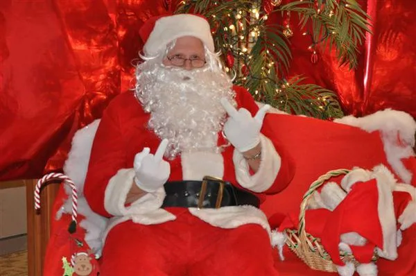 A man dressed as Santa Claus sits on a red chair, making a rude hand gesture with both hands while wearing glasses and a…
