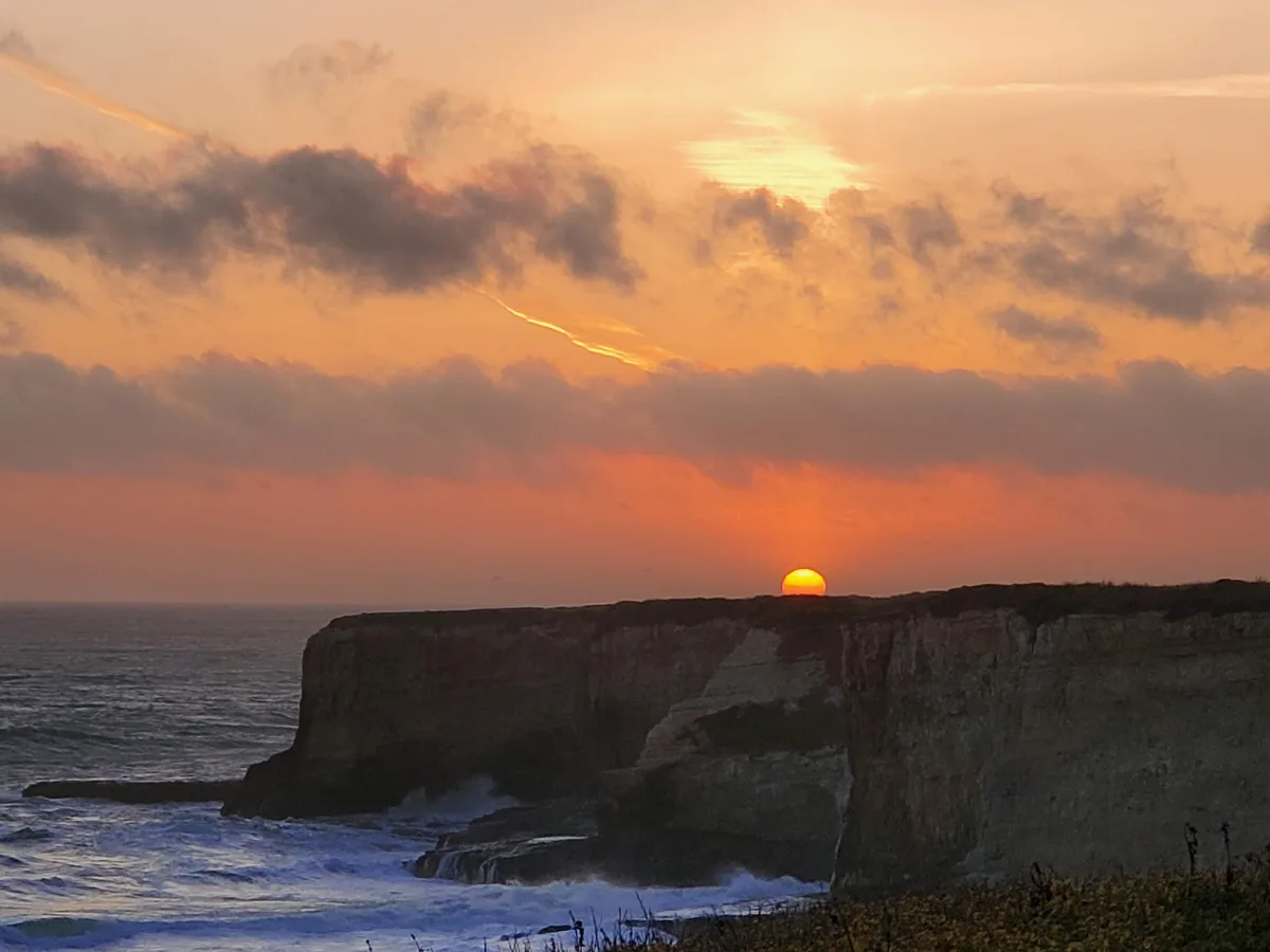 A scenic sunset photograph shows the sun setting directly behind a rugged coastal cliff, casting warm orange and pink light…