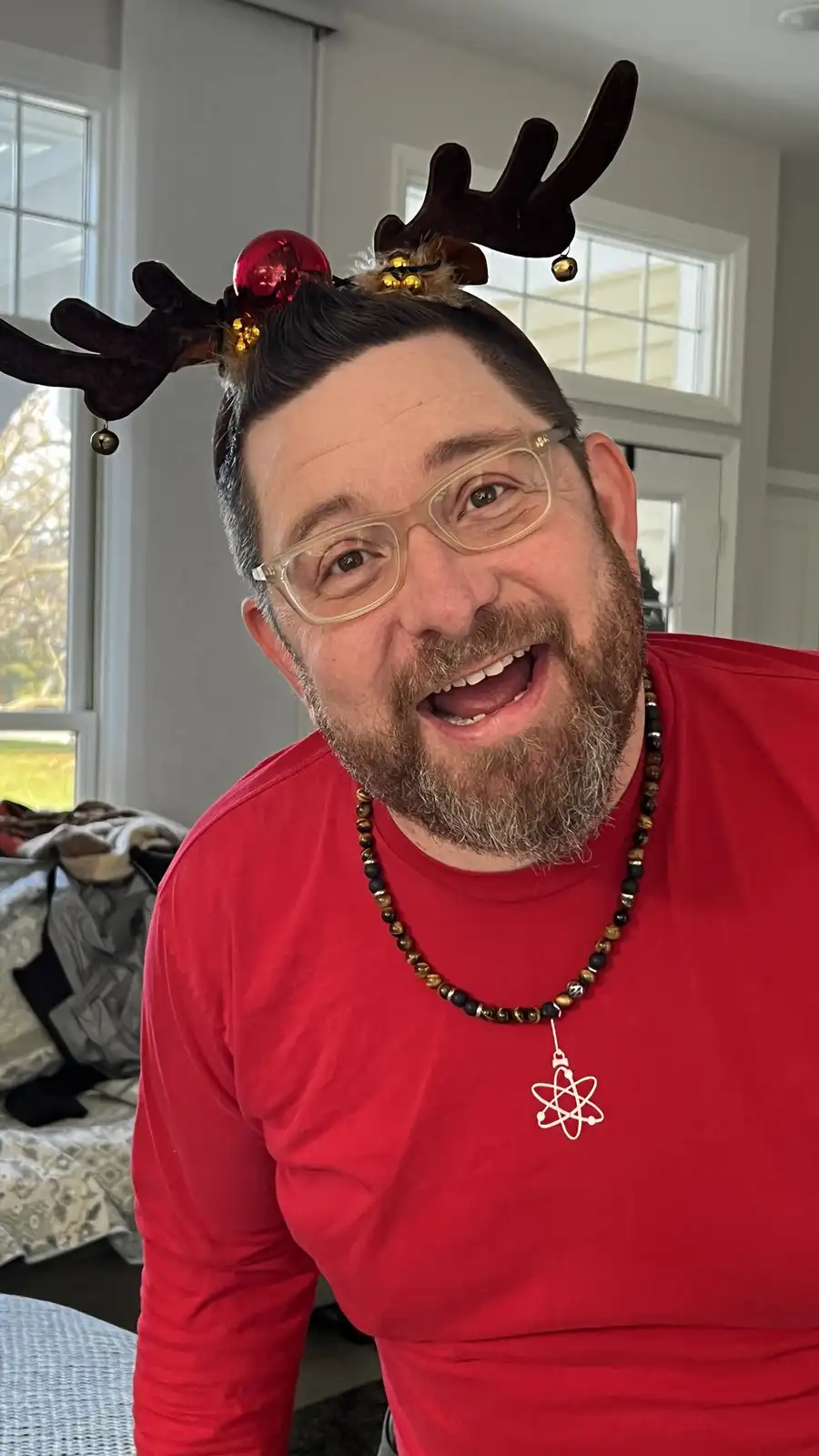 A man with glasses and a beard wears a red shirt and a reindeer antler headband decorated with a red ornament and bells. He…