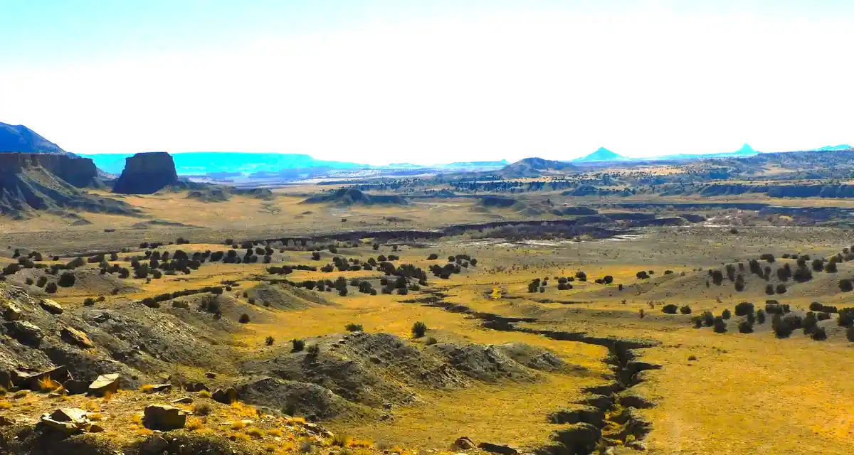A wide, sunlit desert landscape with rolling hills, scattered shrubs, and distant mesas under a bright sky. Overlaid text…