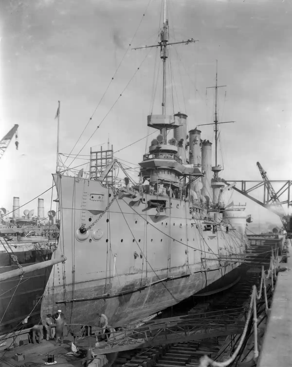 A black-and-white historical photograph shows a large battleship in dry dock, with workers visible on the dock and cranes in…