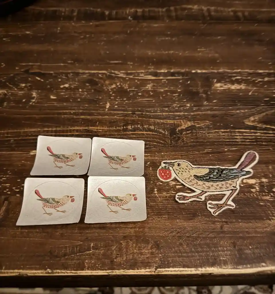 Four identical square stickers of a small bird holding a strawberry are arranged in a grid on a wooden surface. To the…
