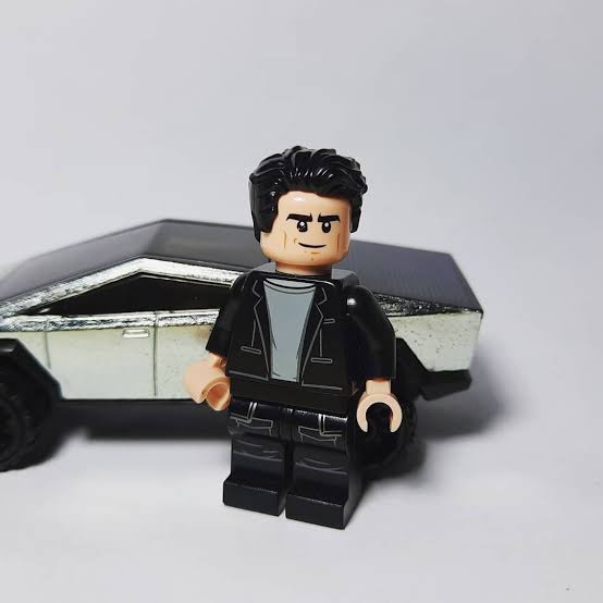 A LEGO minifigure of a man with black hair and a smirk stands in front of a LEGO model of a DeLorean car. The figure wears a…