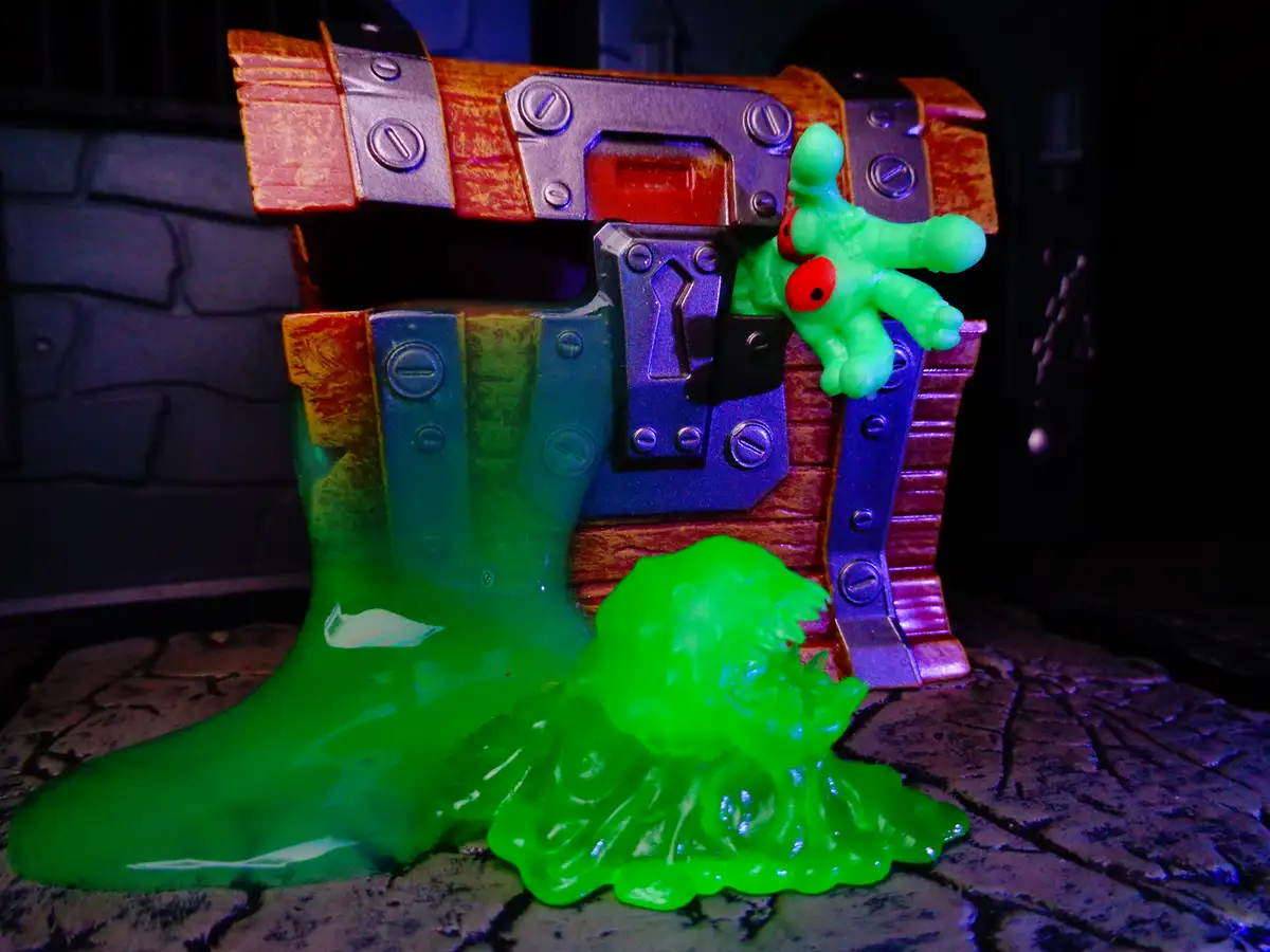 A toy chest with a green, gooey monster figure emerging from it, with green slime pooling on the ground. The monster has red…
