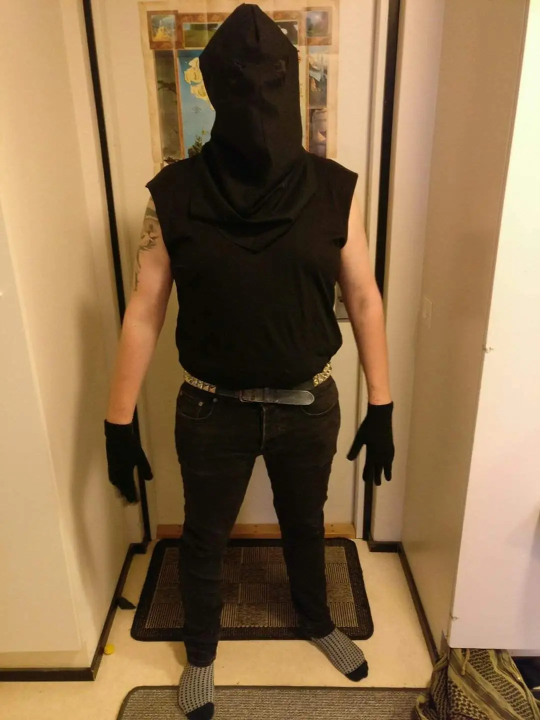 A person stands in a doorway wearing a black balaclava covering their entire head and face, a black sleeveless shirt, black…