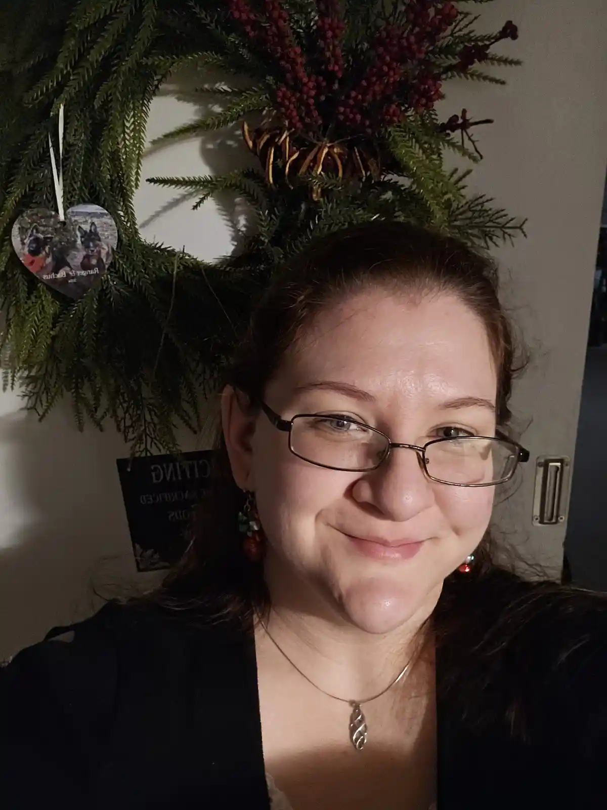 A woman with glasses and a slight smile stands in front of a large green Christmas wreath adorned with red berries and a…