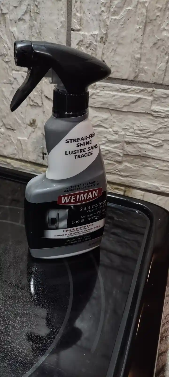 A bottle of Weiman Stainless Steel Cleaner & Polish sits on a black glass stovetop, its label clearly visible against a…