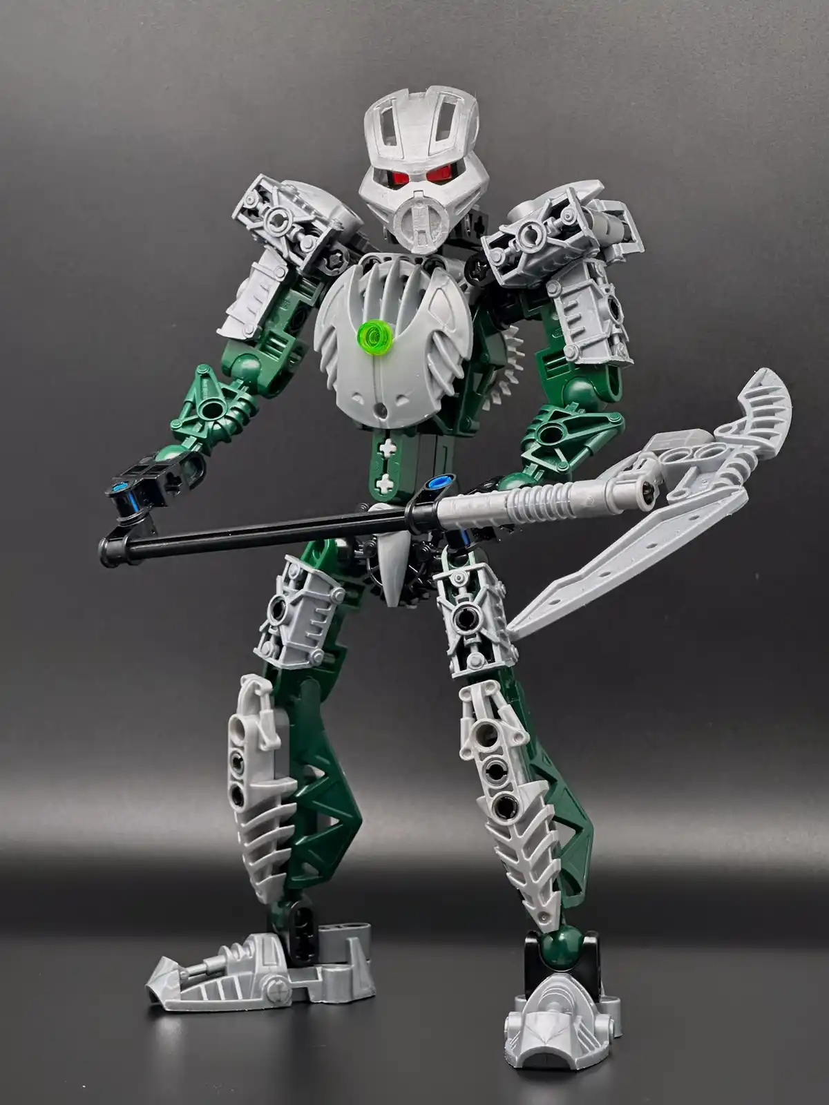 A custom-built Bionicle-style robot figure, primarily silver and green with red eyes, stands in a combat pose holding a…
