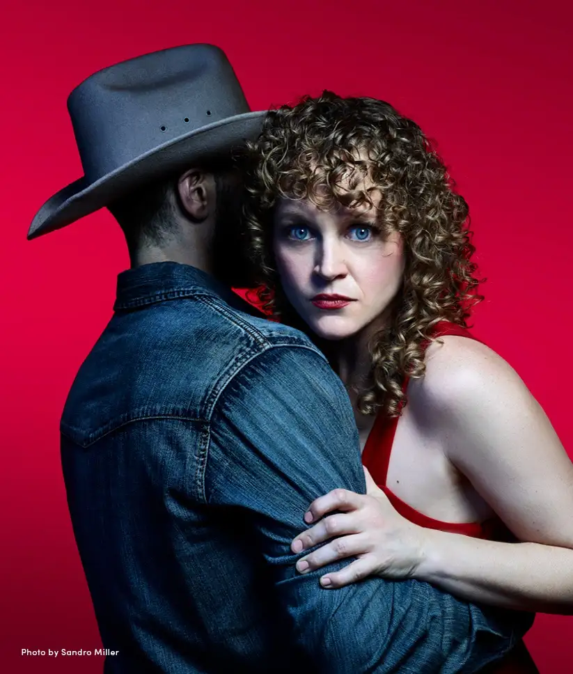 A woman with curly hair and red lipstick looks intensely at the camera while embracing a man from behind; he wears a cowboy…