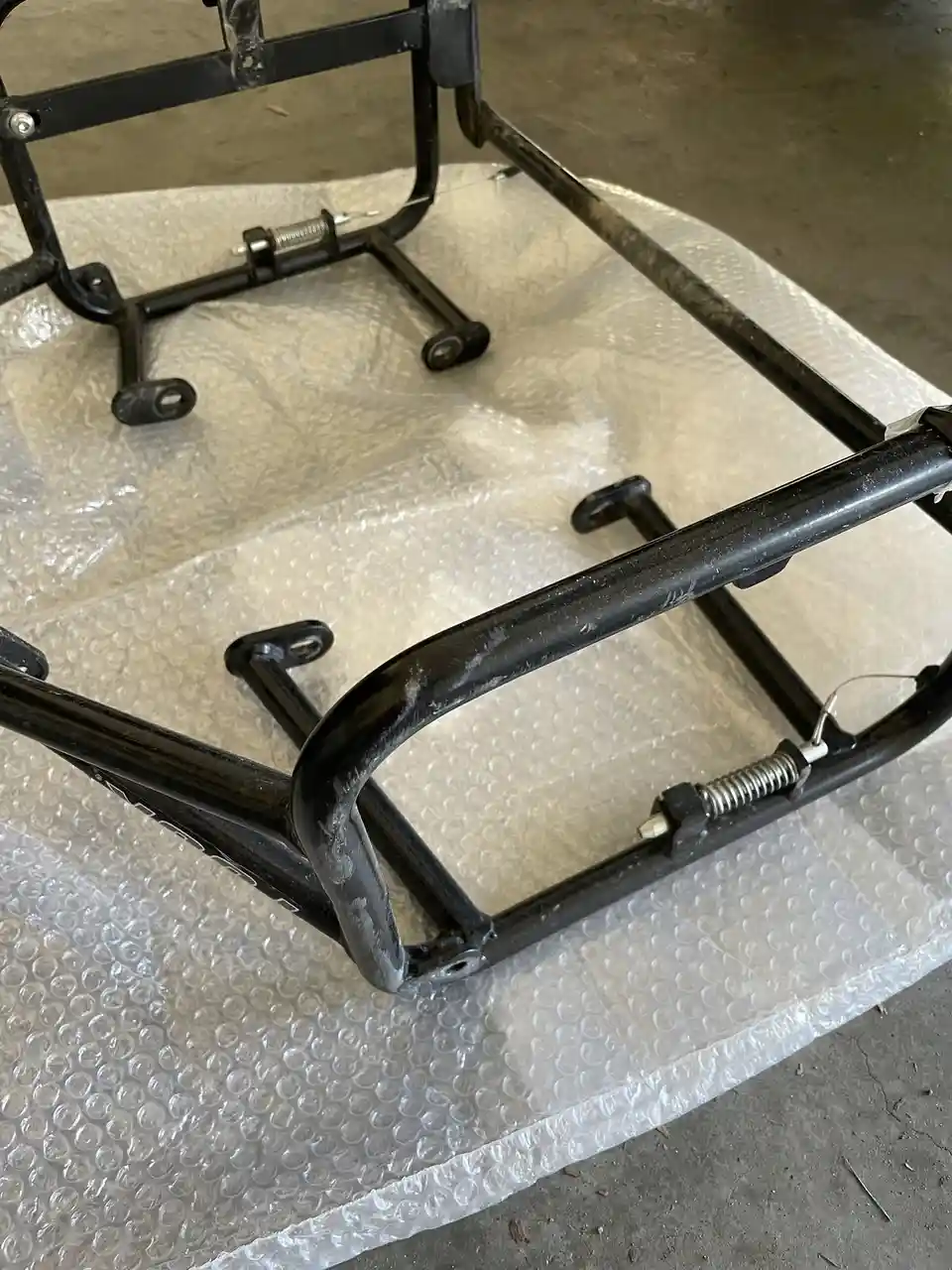 A black metal chair frame, missing all upholstery and components, rests on bubble wrap on a concrete floor. The overlaid…