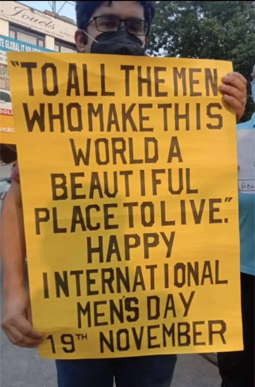 A person wearing glasses and a face mask holds a large yellow sign with bold black text. The sign reads: 'TO ALL THE MEN WHO…