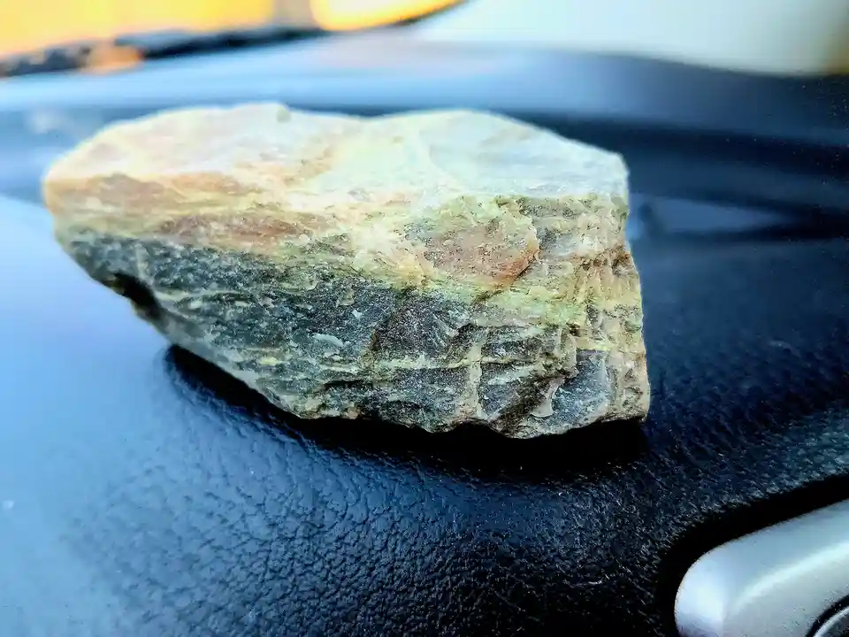 A close-up photo shows a rough, multi-colored rock with layers of beige, green, and gray resting on a black textured…