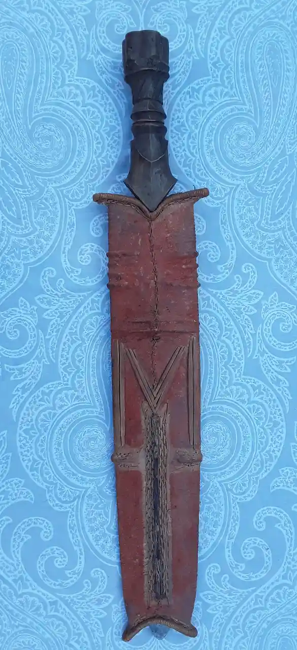 A traditional ceremonial knife with a dark, ornate handle and a red leather sheath featuring woven patterns, displayed…
