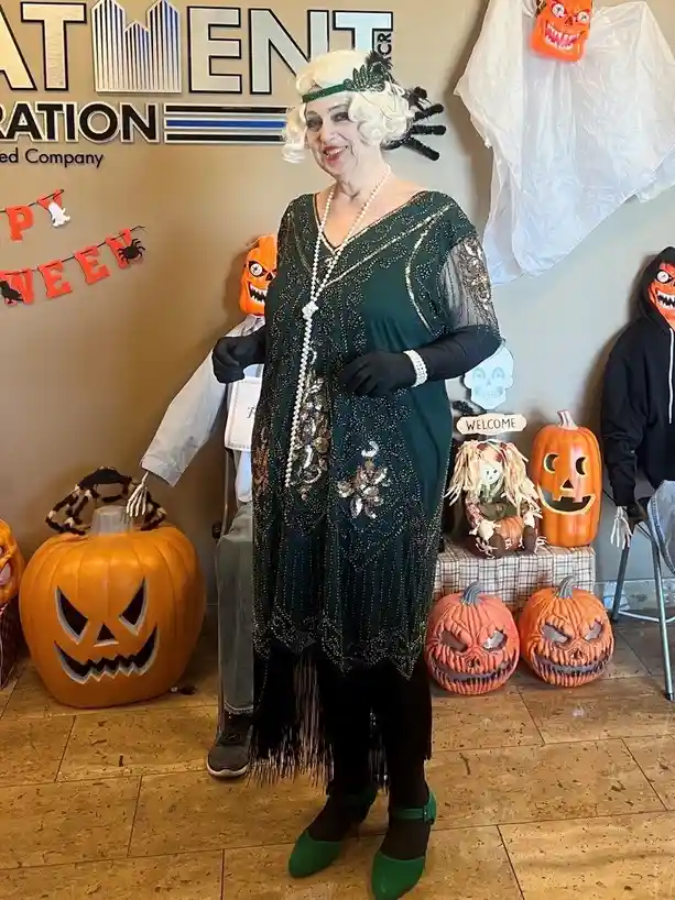 A woman dressed in a 1920s flapper costume with a green beaded dress, pearl necklace, and white wig stands in front of a…