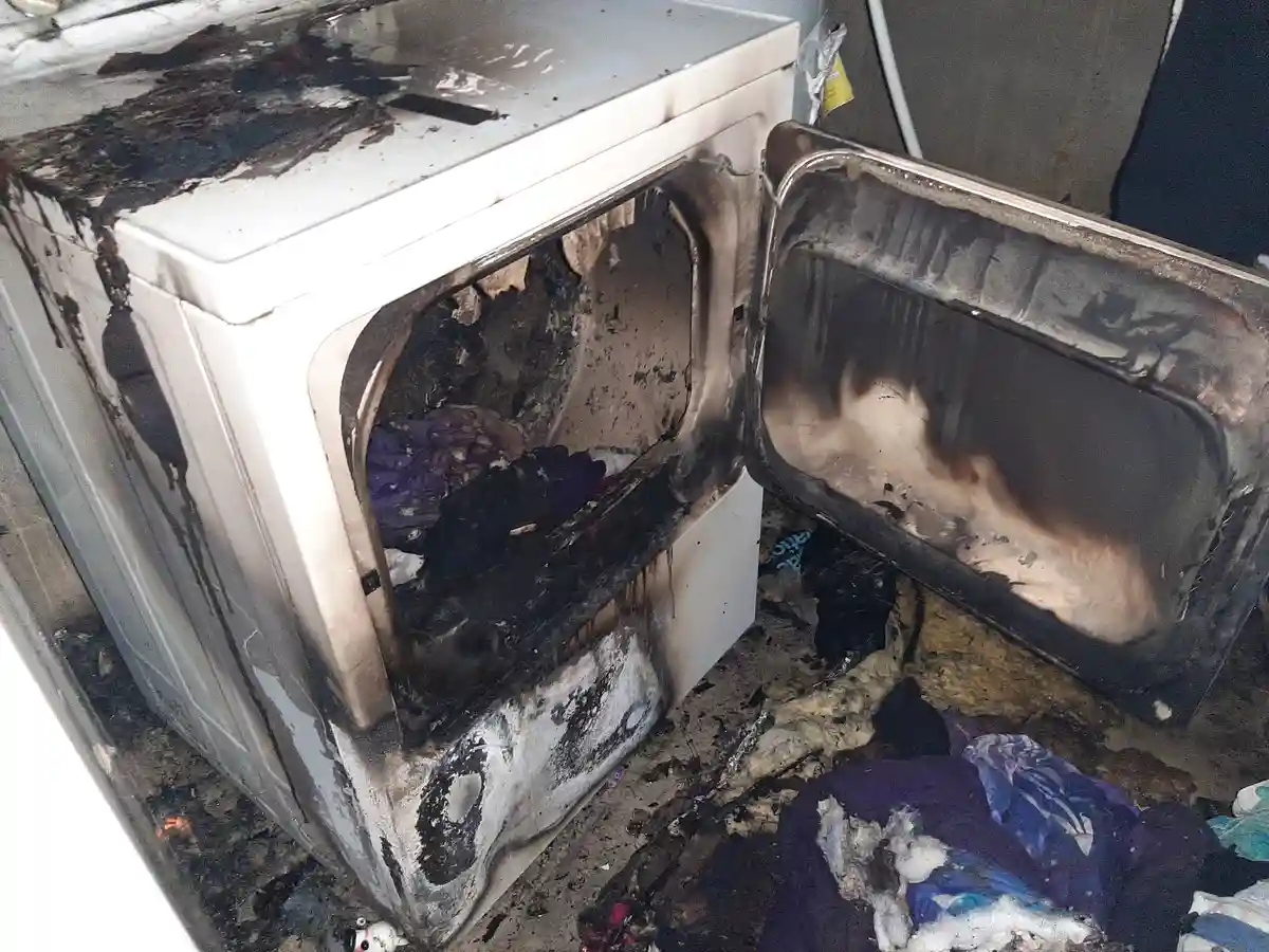 A severely burnt white dryer with its door open, showing charred interior and melted plastic, surrounded by ash and burnt…