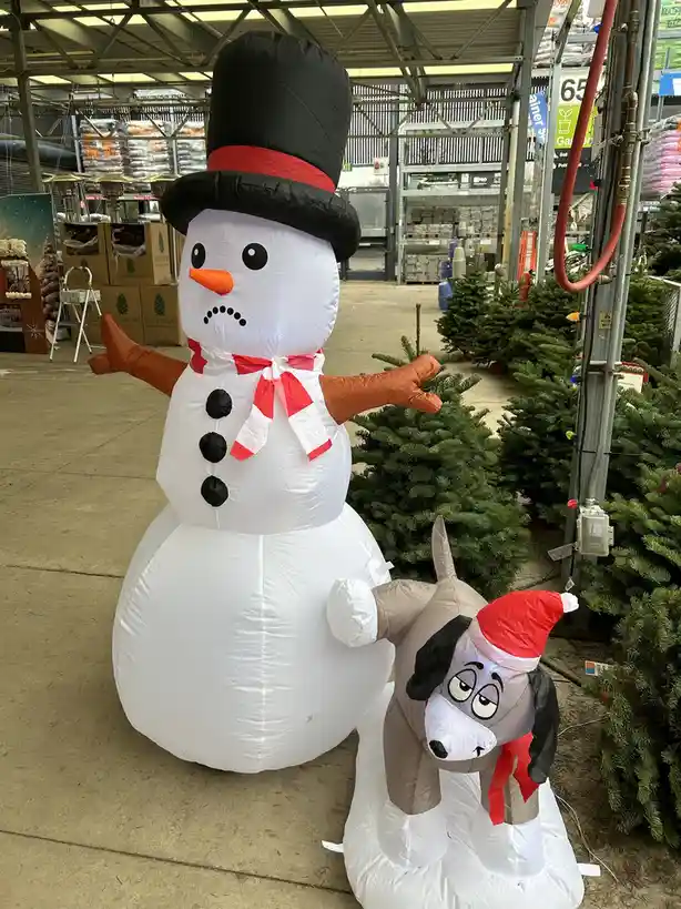 An inflatable snowman wearing a black top hat and red scarf stands next to a smaller inflatable dog wearing a red Santa hat…
