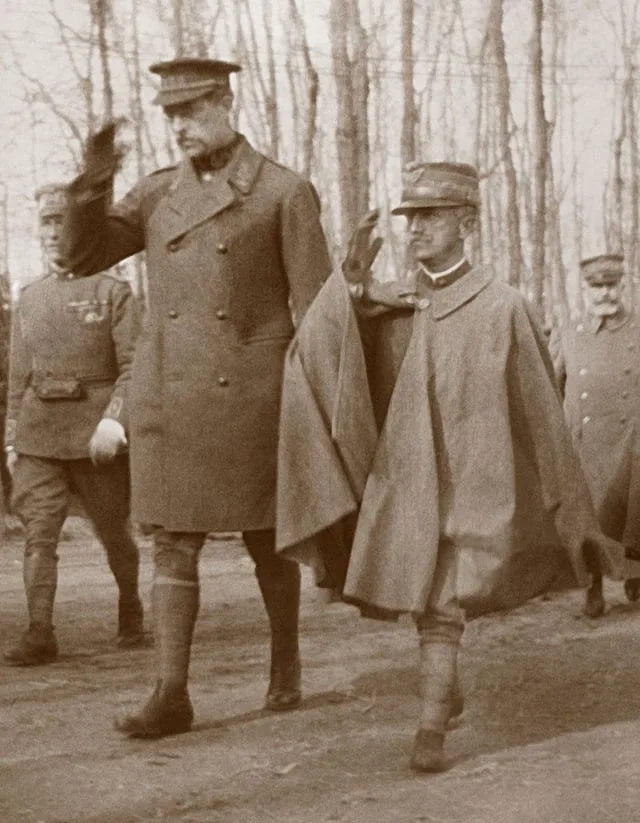 a sepia-toned historical photograph shows several men in military uniforms walking outdoors in a wooded area. The central…