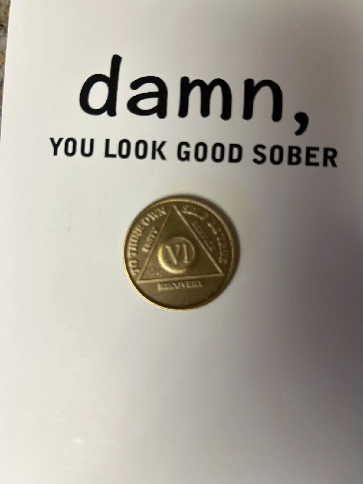A white surface displays the text 'damn, YOU LOOK GOOD SOBER' in black font above a gold-colored recovery token. The token…