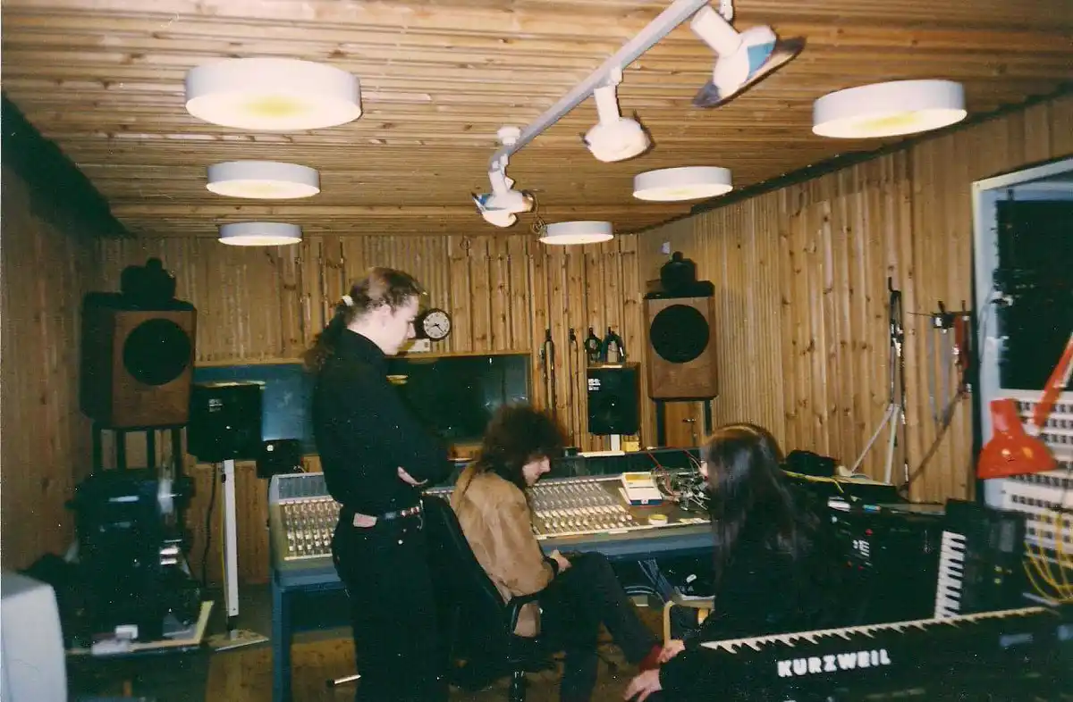 A grainy photo shows three people in a wood-paneled recording studio with vintage equipment, including a mixing board…