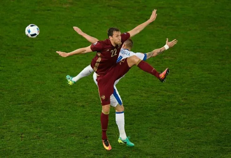Two soccer players are mid-air, tangled in a chaotic collision during a match. The player in the maroon jersey with number…