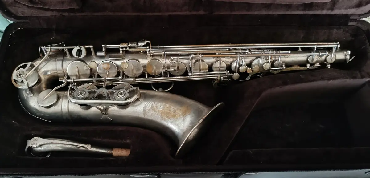 A silver saxophone with visible wear and tarnish rests inside a dark, plush-lined case, with its mouthpiece and ligature…
