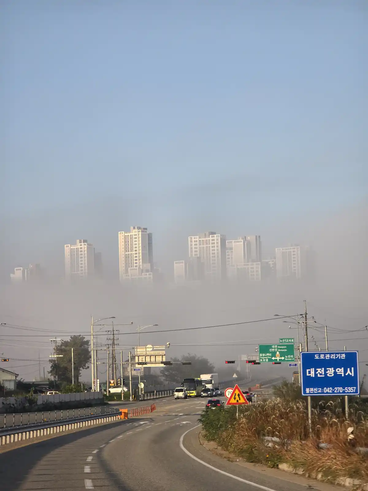 A road scene in Daejeon, South Korea, where a thick layer of fog obscures the city skyline in the background. Visible are…