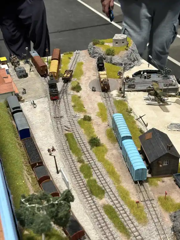 An overhead view of a highly detailed model train layout featuring multiple tracks, miniature trains (including a steam…