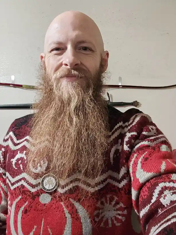 A bald man with a long, full beard and mustache smiles at the camera while wearing a red and white patterned sweater. Behind…
