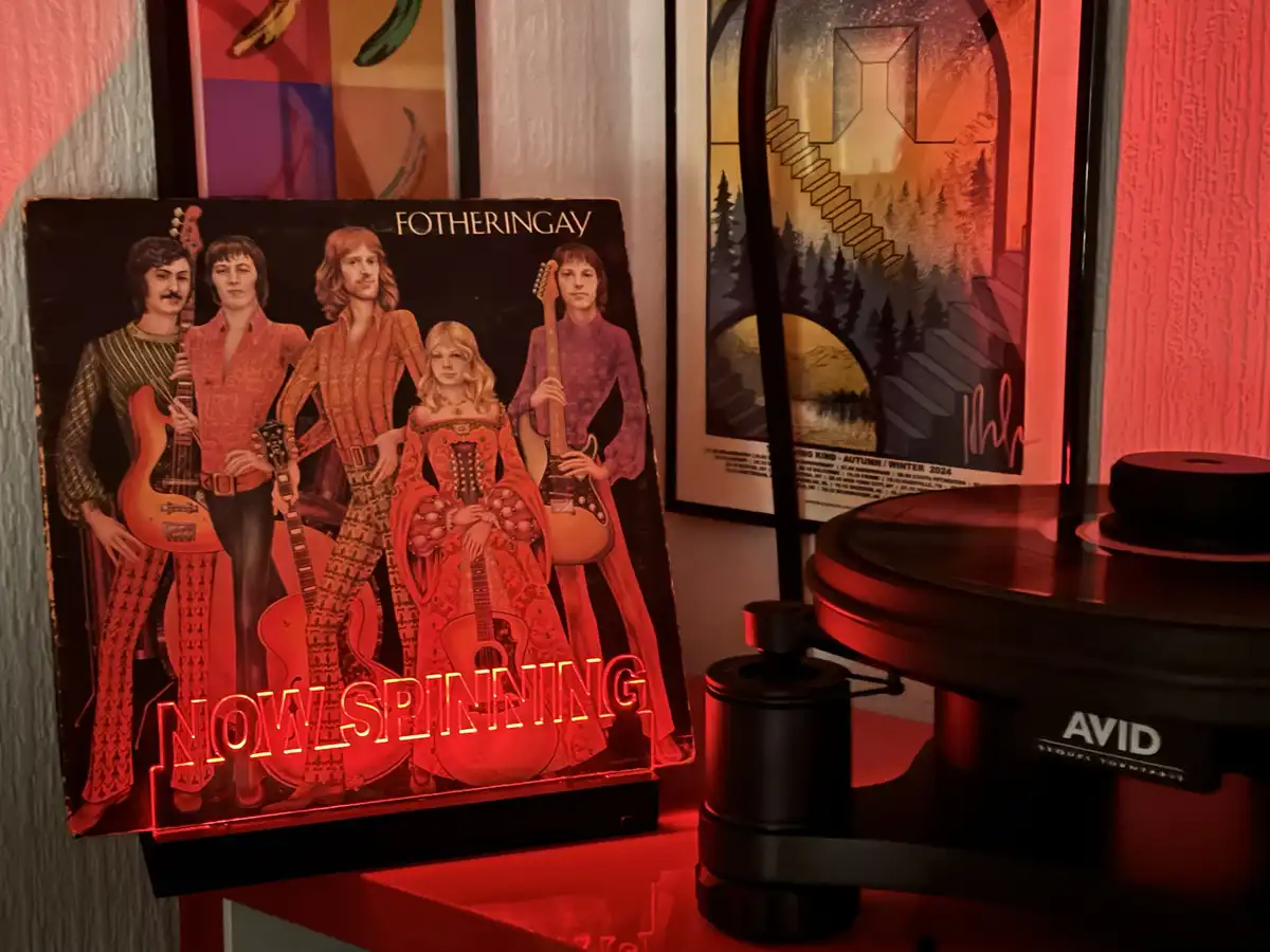 A vinyl record album cover for the band Fotheringay is displayed upright on a shelf next to a black AVID turntable, both…
