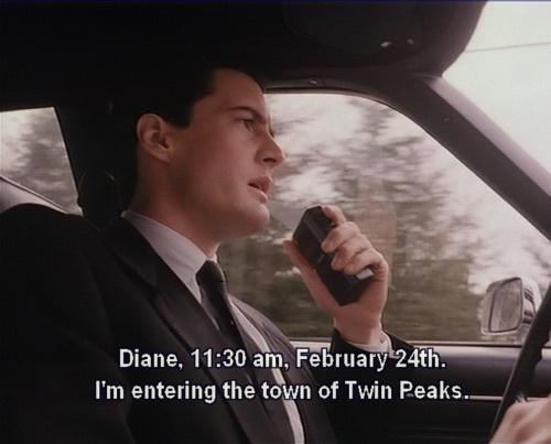 A man in a suit and tie is driving a car while holding a walkie-talkie to his mouth. The image is from the TV show Twin…