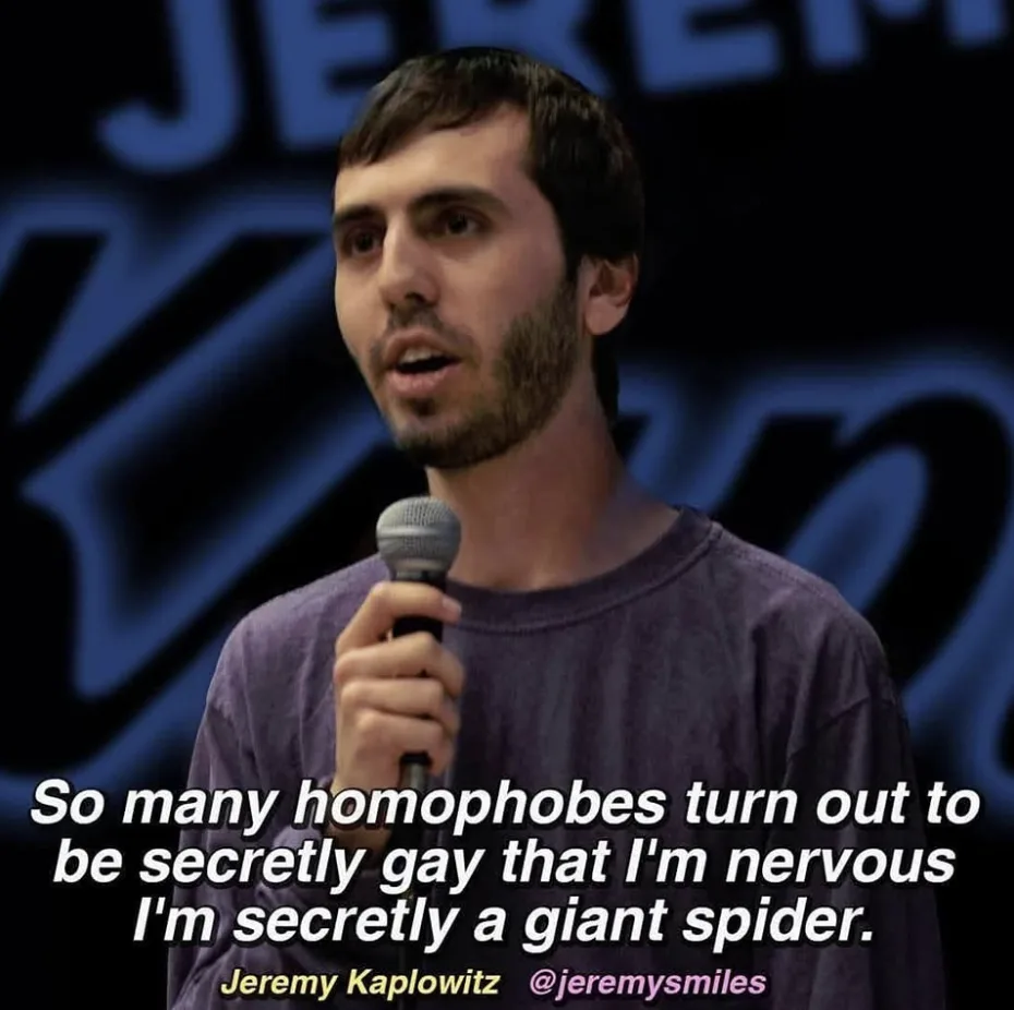 A man with short dark hair and a beard, wearing a purple t-shirt, holds a microphone while speaking. Behind him is a dark…