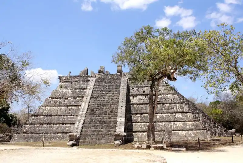 A large, ancient stone Mayan pyramid with a wide staircase under a bright blue sky, partially framed by green trees…