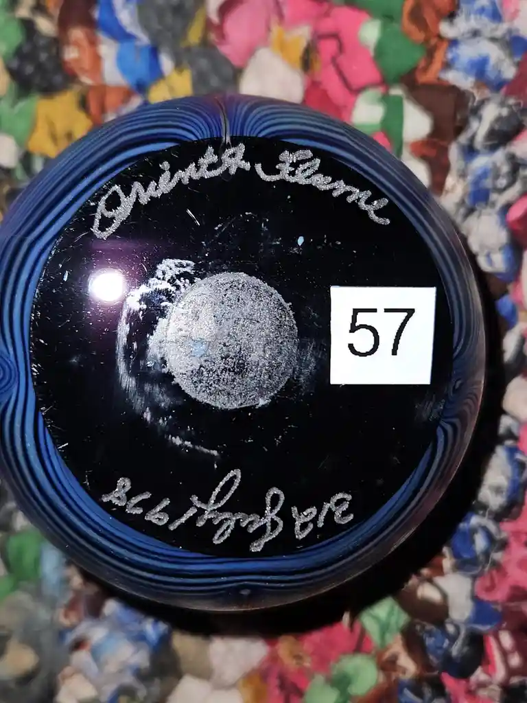 A close-up of a black bowling ball with blue concentric rings, showing a silver glittery signature 'Quinta Elena' at the…