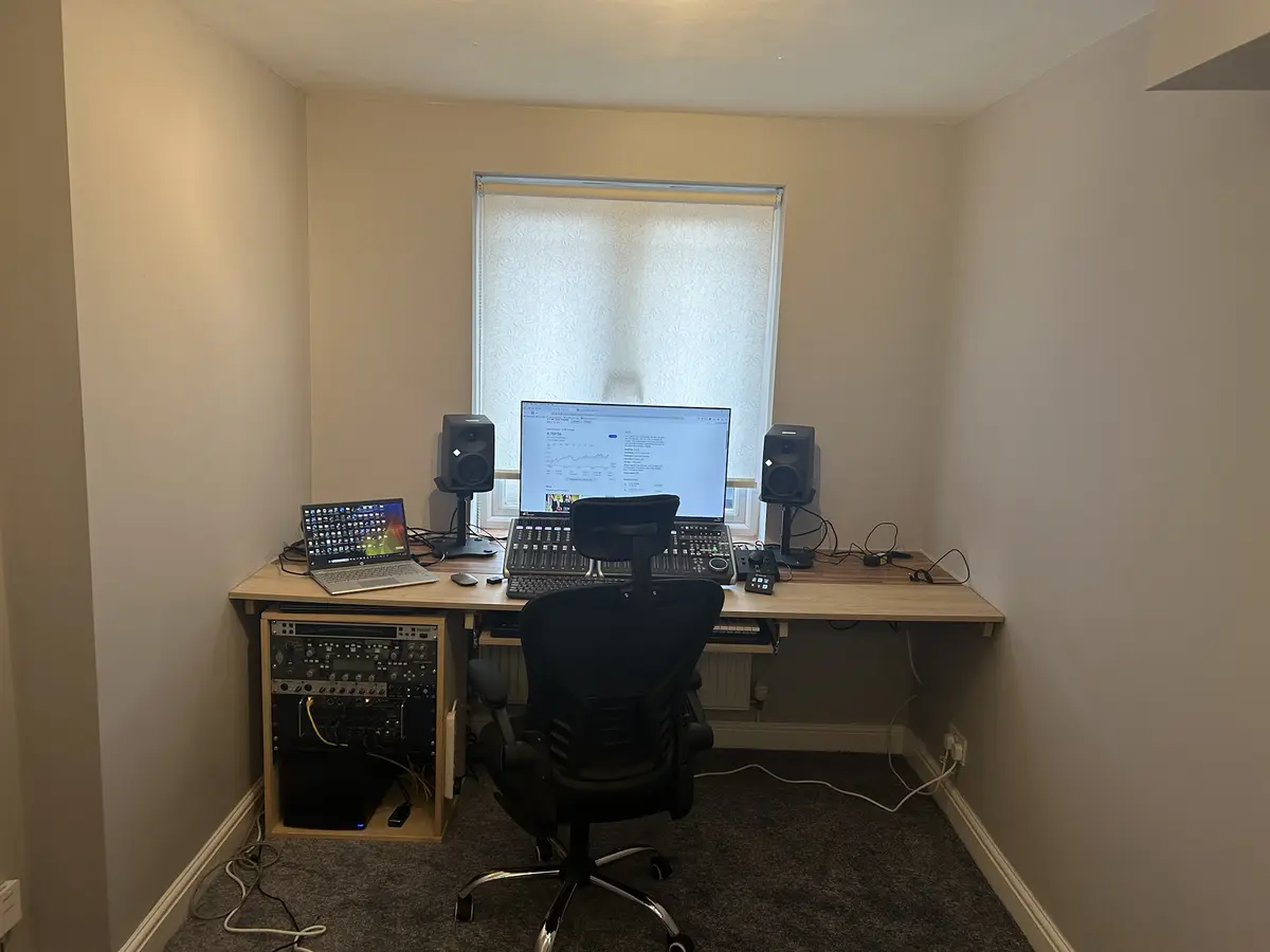 A home studio setup with a desk, black ergonomic chair, laptop, large monitor displaying a music production interface, and…