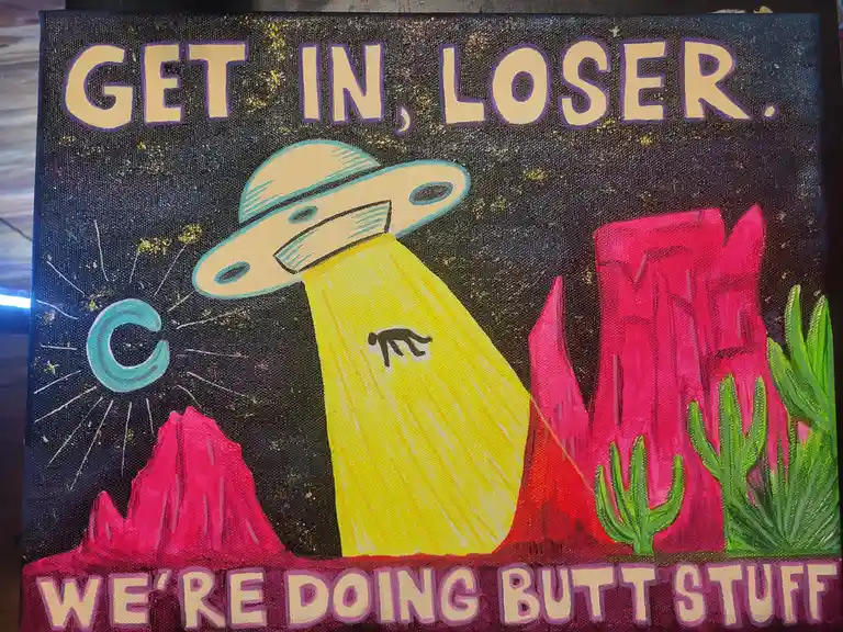 A hand-painted canvas depicts a cartoon UFO hovering over a desert with pink rock formations and green cacti, emitting a…
