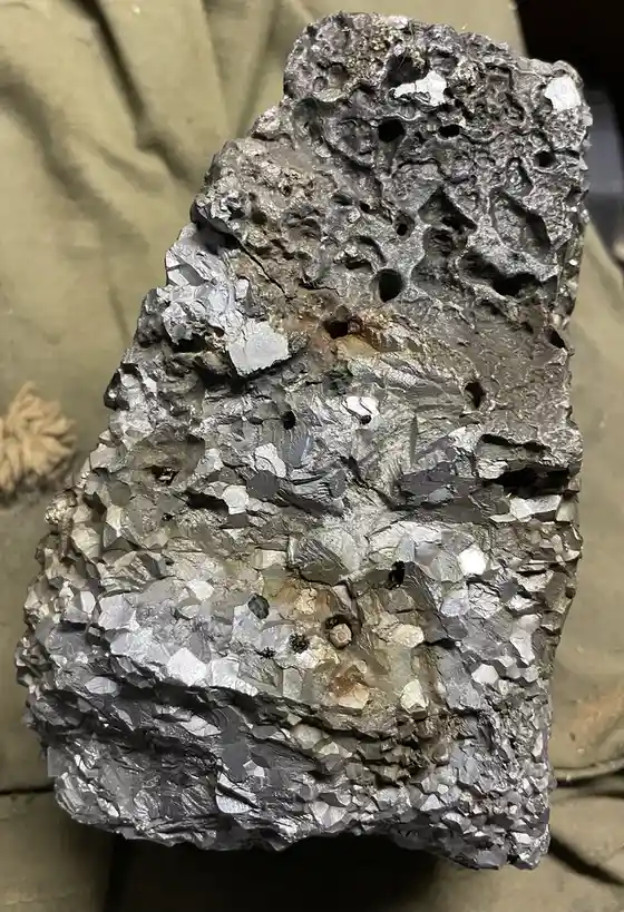 A close-up photo shows a rough, irregularly shaped rock with a metallic, silver-gray surface featuring many small, shiny…