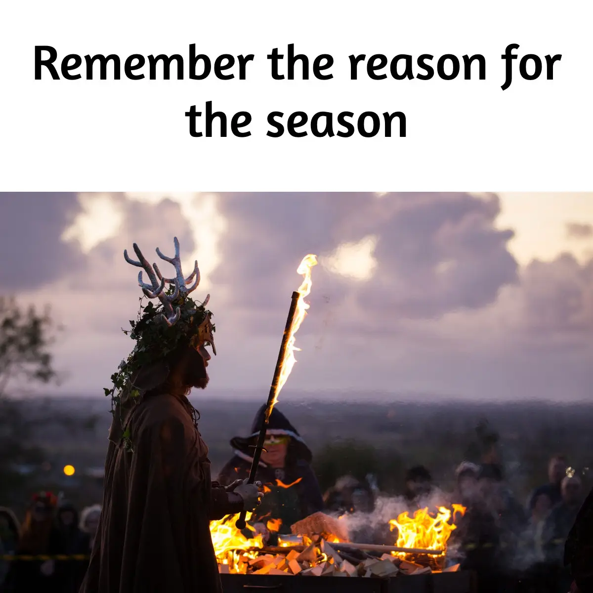 A person in a costume with antlers and foliage on their head holds a flaming torch while standing near a large bonfire at…