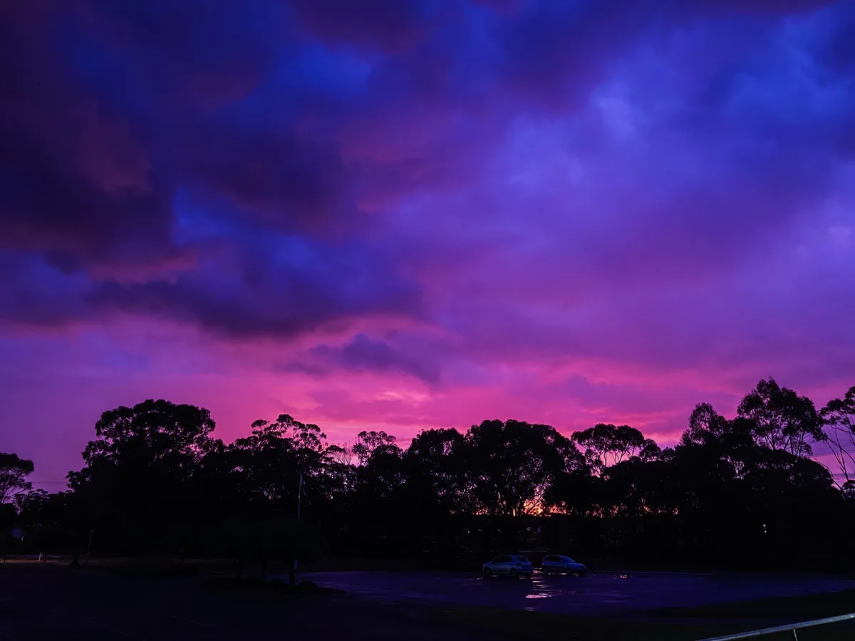 A dramatic sky filled with swirling clouds in deep purple, blue, and pink hues, silhouetting trees and two cars in a parking…