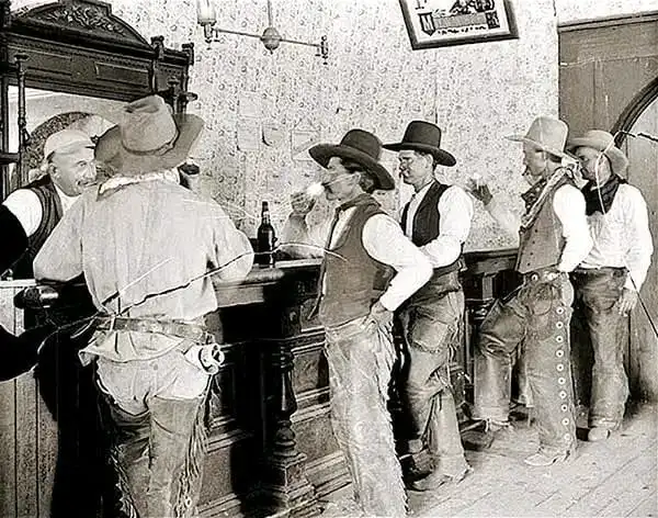 A black-and-white historical photo shows several men in cowboy attire gathered around a saloon bar, some holding drinks. One…