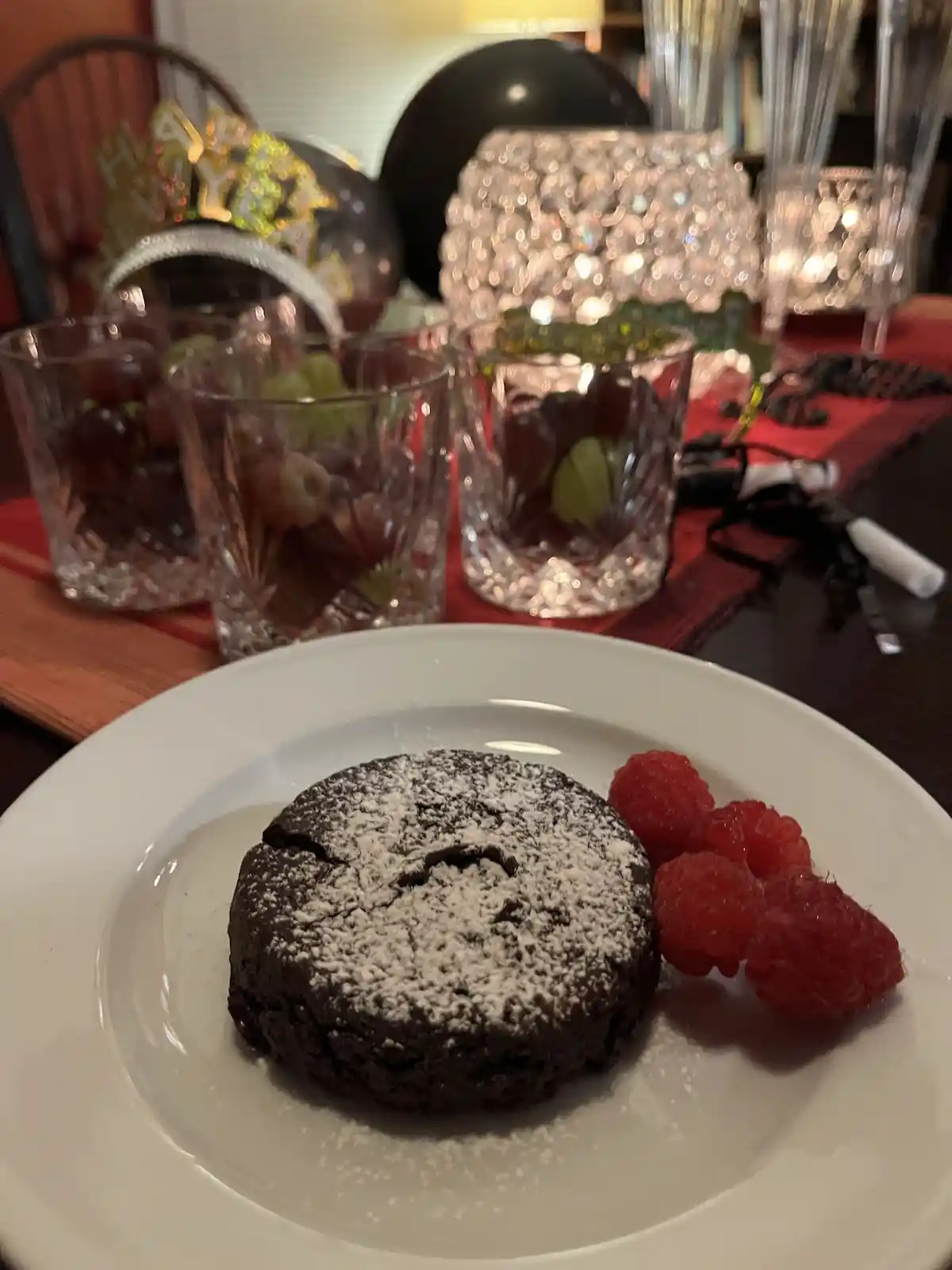 A close-up photo shows a small chocolate lava cake dusted with powdered sugar on a white plate, next to fresh raspberries…