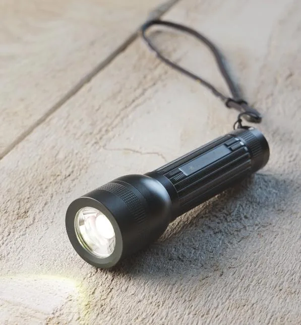 A black, textured flashlight with a lanyard lies on a rough, light-colored stone surface, its beam shining brightly on the…