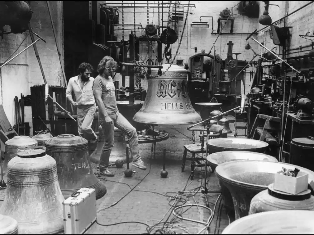 A black-and-white photo shows two men in a bell foundry surrounded by large bronze bells. One man sits on a bell while the…