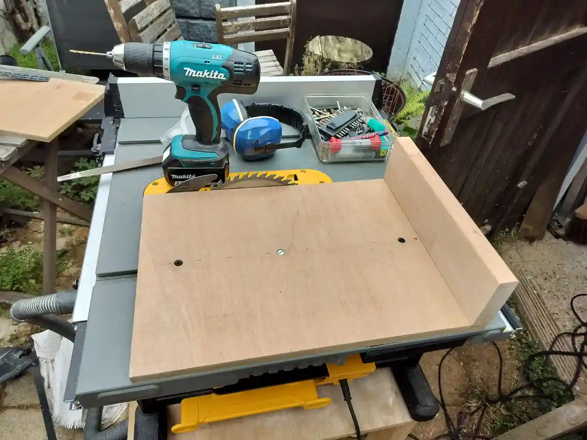 A woodworking setup with a Makita drill, ear protection, and tools on a table saw, featuring a wooden board with drilled…