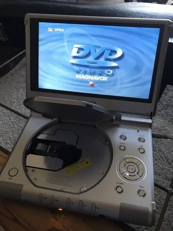 A silver Magnavox portable DVD player with its lid open, revealing an empty disc tray. The screen displays the 'DVD VIDEO'…