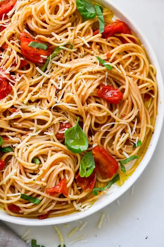 a close-up photo of a white bowl filled with spaghetti tossed in tomato sauce, garnished with roasted cherry tomatoes, fresh…