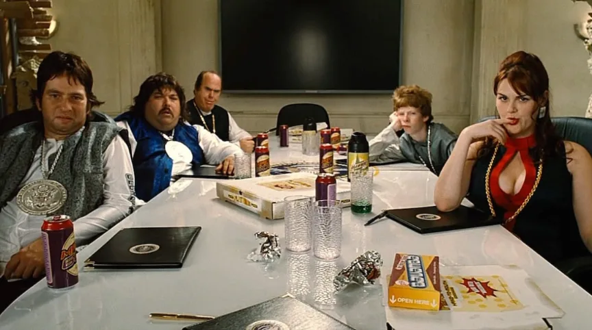 A group of people sit around a conference table with pizza boxes, soda cans, and snacks. The woman on the right, wearing a…